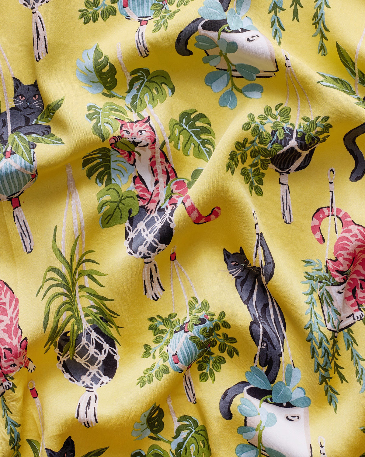 A person wears the PRINTFRESH - Macrame Mischief Satin Wildest Dream Pajama Set in Sunshine, featuring a vibrant cat and hanging plant pattern; their face is partially out of frame as they gently hold the shirt near the collar.
