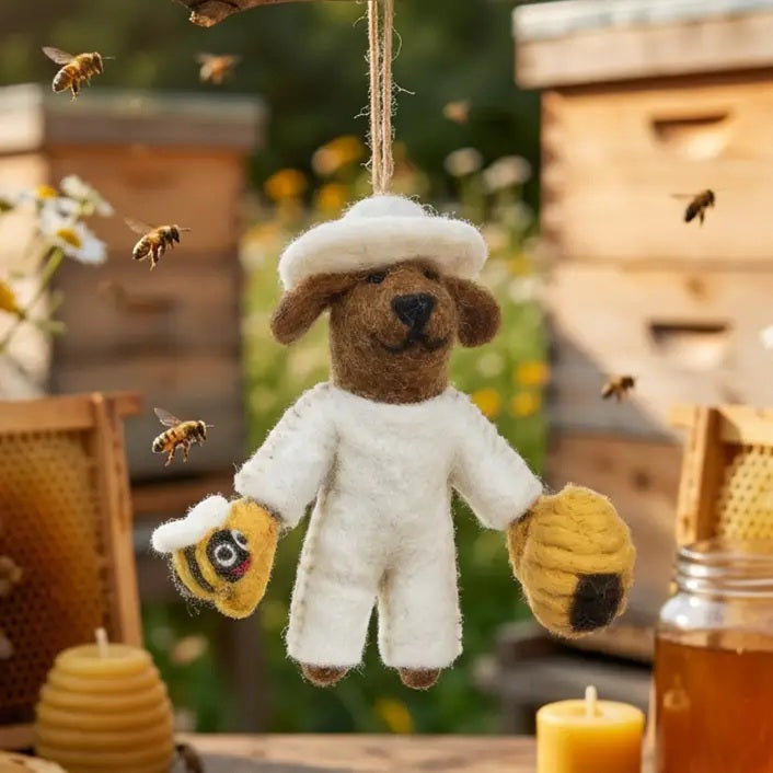 BUMBLE THE BEEKEEPER ORNAMENT is a needle felted dog dressed in a white beekeeper suit, holding a small bee in one hand and a beehive in the other.