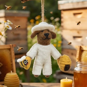 BUMBLE THE BEEKEEPER ORNAMENT is a needle felted dog dressed in a white beekeeper suit, holding a small bee in one hand and a beehive in the other.