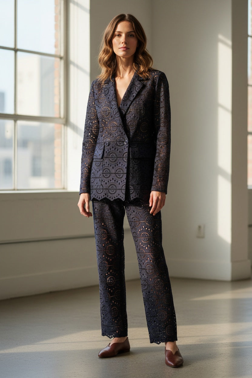 A woman with long dark hair wears the SFIZIO Double Breasted Broderi Anglaise Jacket by SFIZIO, a navy blue lace pantsuit with a geometric eyelet pattern, standing before yellow curtains.