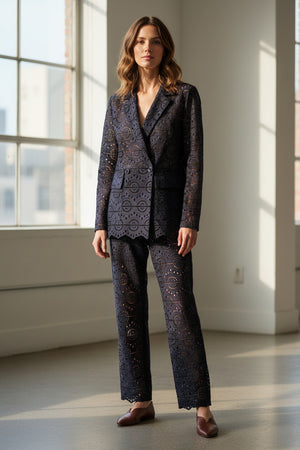 A woman with long dark hair wears the SFIZIO Double Breasted Broderi Anglaise Jacket by SFIZIO, a navy blue lace pantsuit with a geometric eyelet pattern, standing before yellow curtains.