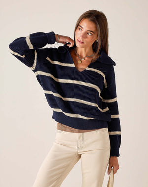 Wearing the MERSEA Camden Sailor Sweater in Navy Cream—adorned with classic white stripes—a woman stands in a sunlit room with expansive windows, smiling as she pairs the look with cream high-waisted pants, effortlessly channeling coastal Maine charm.