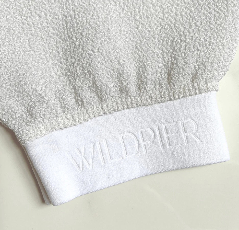 The WILDPIER DEEP EXFOLIATING GLOVE is white with a slightly rounded top and elastic wristband, designed to reveal radiant skin. It's shown against a plain white background.