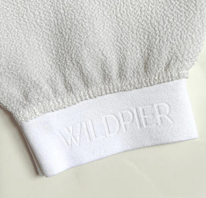 The WILDPIER DEEP EXFOLIATING GLOVE is white with a slightly rounded top and elastic wristband, designed to reveal radiant skin. It's shown against a plain white background.