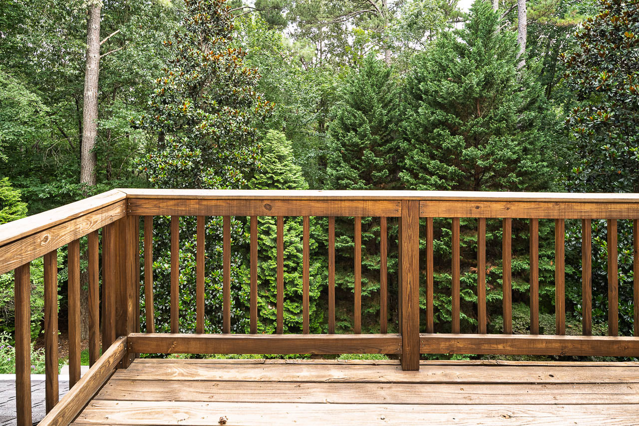 A wooden deck with a railing overlooks dense, green trees and foliage in a forested area on a bright day.
