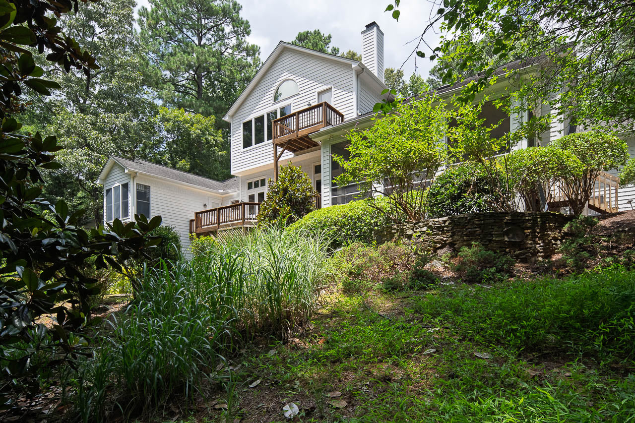 A two-story white house with a small balcony and large windows, surrounded by lush greenery, tall trees, shrubs, and a grassy, slightly sloped yard. The scene is bright and sunny.
