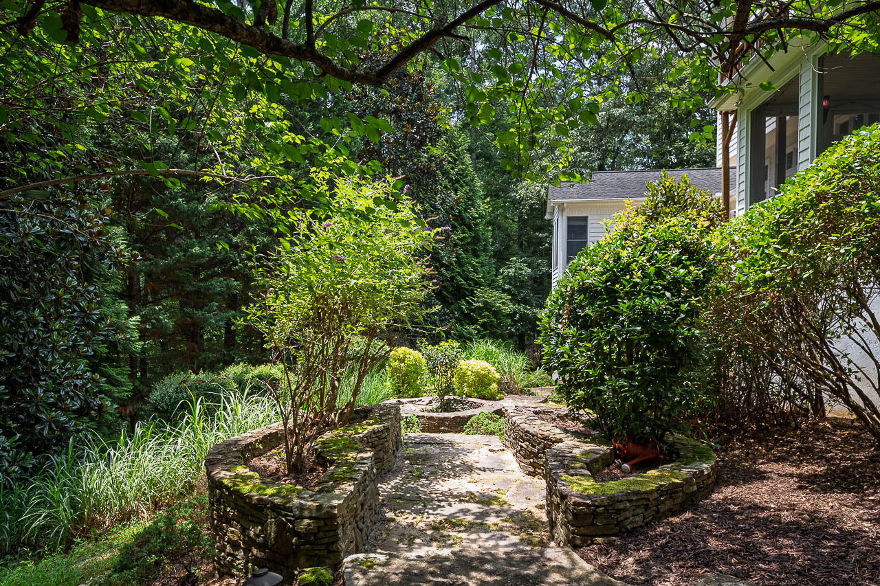 A stone path lined with low stone walls curves through a lush, green garden next to a house, shaded by overhanging tree branches and surrounded by various bushes and plants.