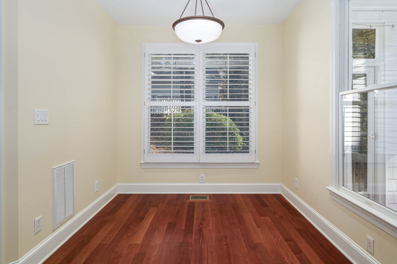 A small, empty room with light yellow walls, a large window with white shutters, reddish-brown hardwood floors, and a round ceiling light fixture. Sunlight streams in through the window.