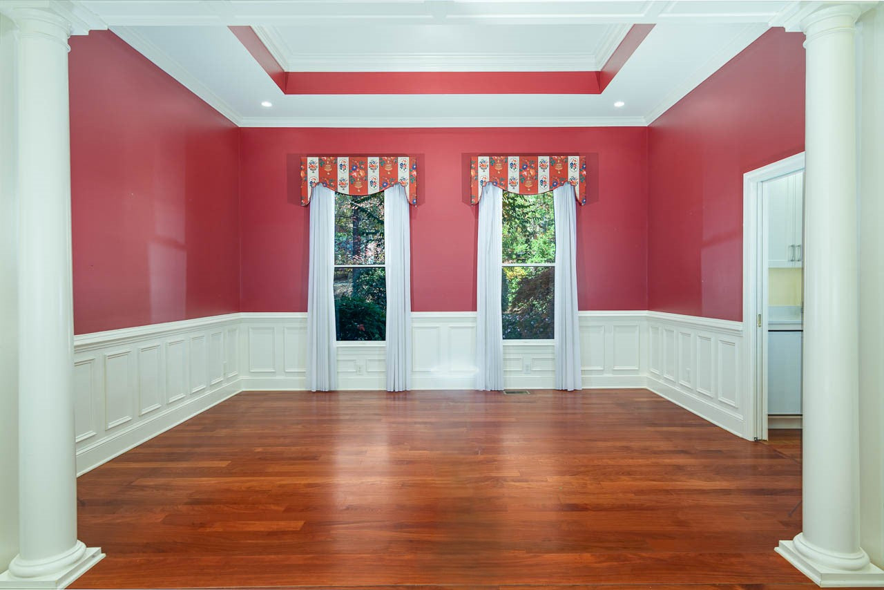 A spacious room with red walls, white wainscoting, two tall windows with white curtains and patterned valances, hardwood floors, and white columns framing the entrance.