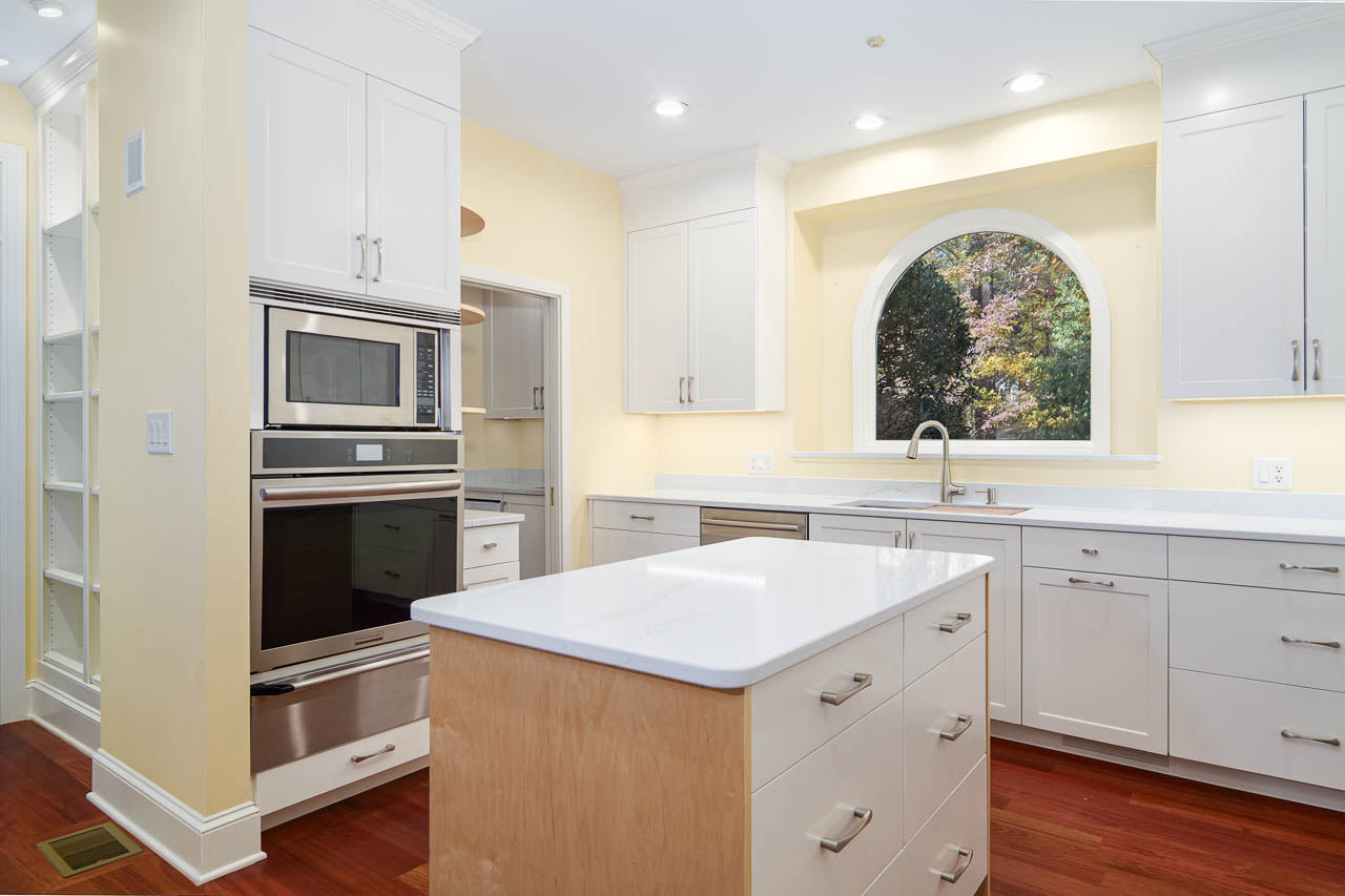 Modern kitchen with white cabinets, stainless steel appliances, a center island, and a large arched window above the sink. The space has wood floors and recessed lighting, creating a bright and clean atmosphere.