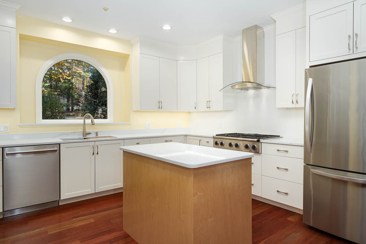 Modern kitchen with white cabinets, stainless steel appliances, a central island, a large arched window above the sink, and wooden floors. The walls are painted light yellow, and the kitchen is well-lit.