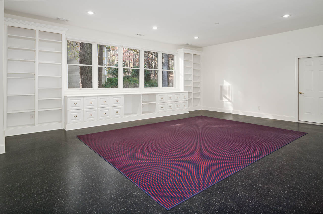 A spacious, bright room with large windows, built-in white shelves and cabinets, black flooring, and a large purple area rug in the center. A closed white door is on the right wall.