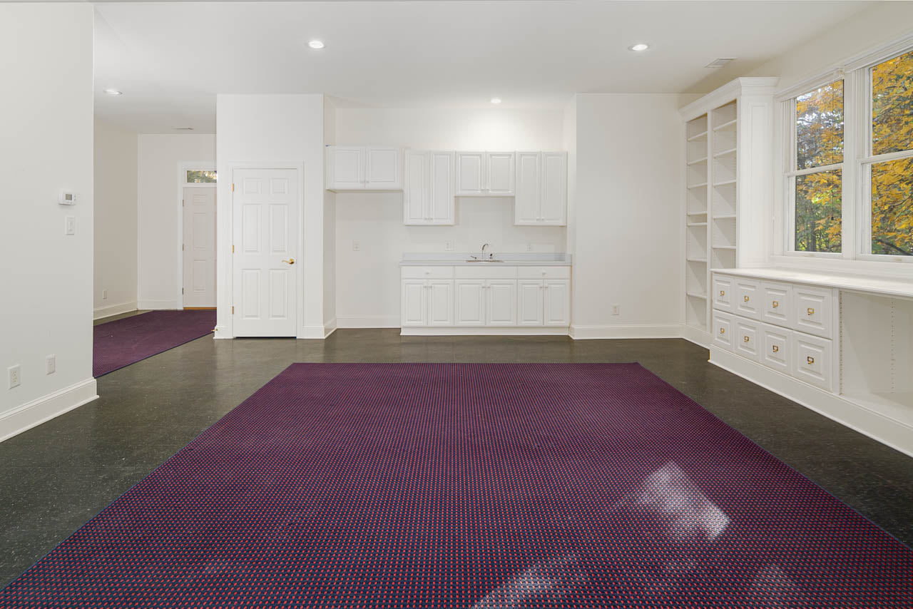 A spacious, empty room with white built-in cabinets, a small sink, open shelving, and a large window. The floor is dark with a large purple area rug in the center. Walls and ceiling are white.