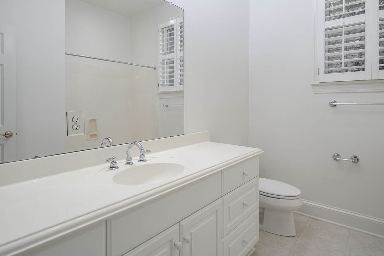 A white bathroom with a large mirror, built-in sink, cabinets, a toilet, and a window with shutters. The walls and countertop are white, creating a clean, bright appearance.