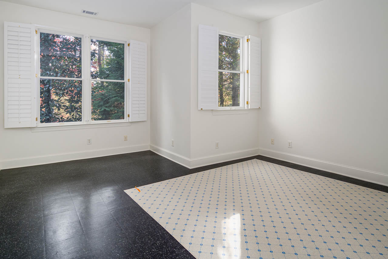 Bright, empty room with white walls, two large windows with white shutters, black tile flooring, and a cream area rug with a blue diamond pattern. Sunlight filters in, highlighting the clean, minimalist space.