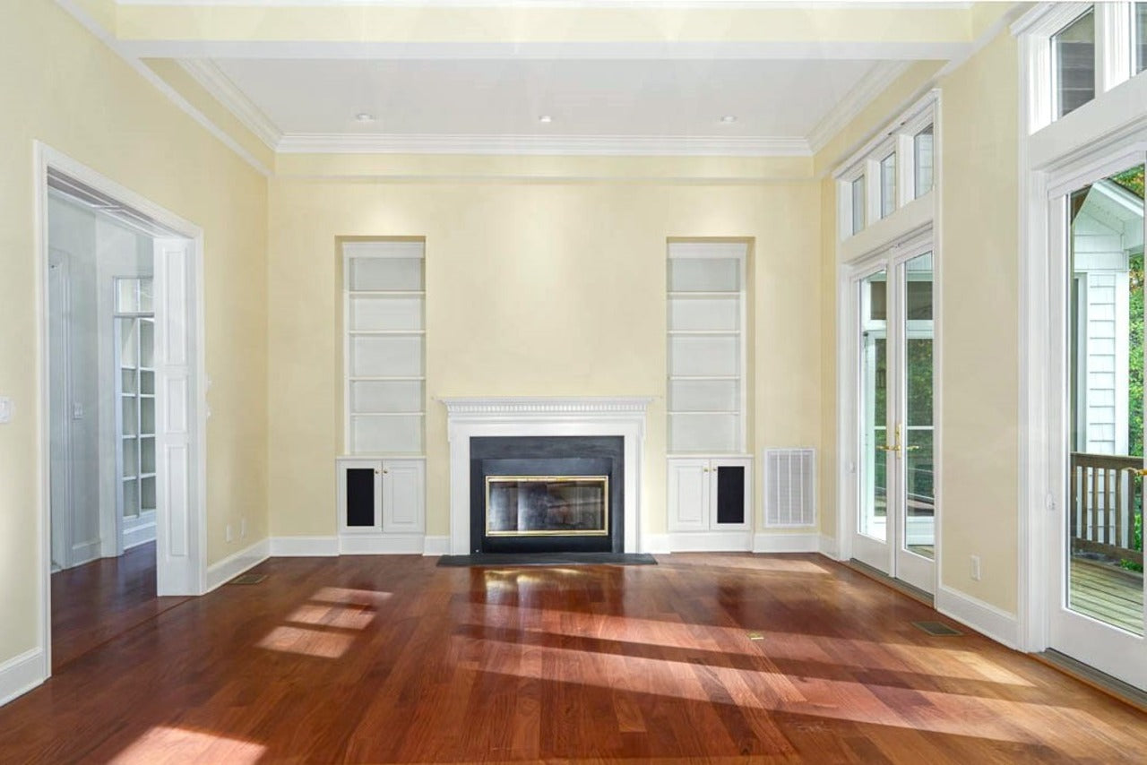 Bright, empty living room with hardwood floors, cream walls, white fireplace, built-in shelves, tall windows, and glass doors opening to an outdoor area. Sunlight streams across the floor.