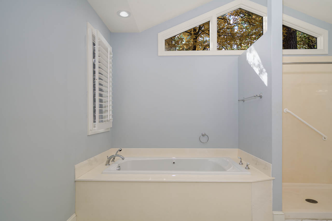 A bathroom with a white bathtub, light blue walls, a window with shutters, a separate shower area, and a high ceiling with triangular windows showing tree branches outside.