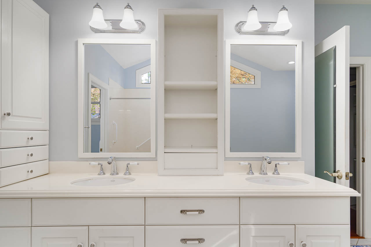 A bright bathroom with double sinks, white cabinets, two mirrors, a central open shelf, and overhead light fixtures. A door is open to the right, revealing a shower area with grab bars.