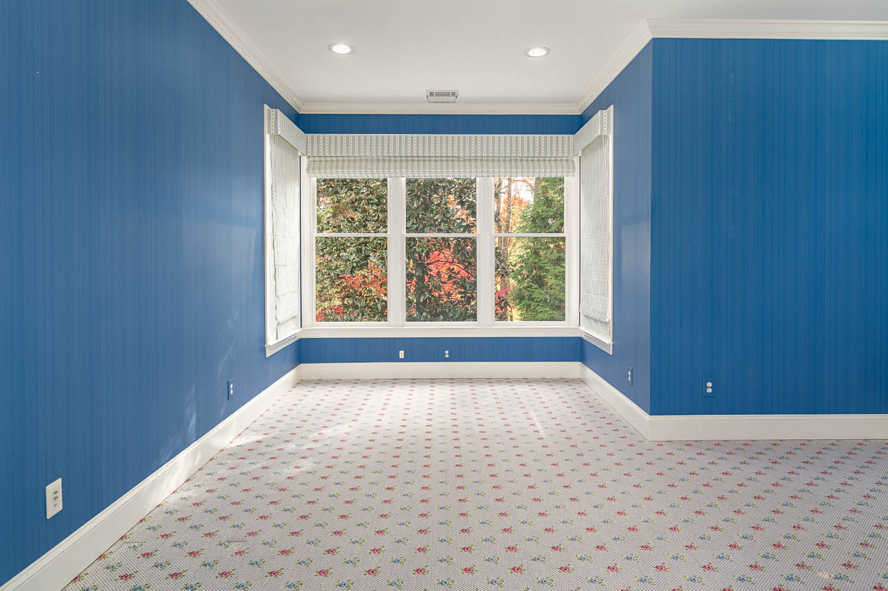 Bright, empty room with blue walls, large windows showing trees outside, and patterned carpet with small red and green floral motifs. White trim and ceiling with recessed lighting add to the airy feel.