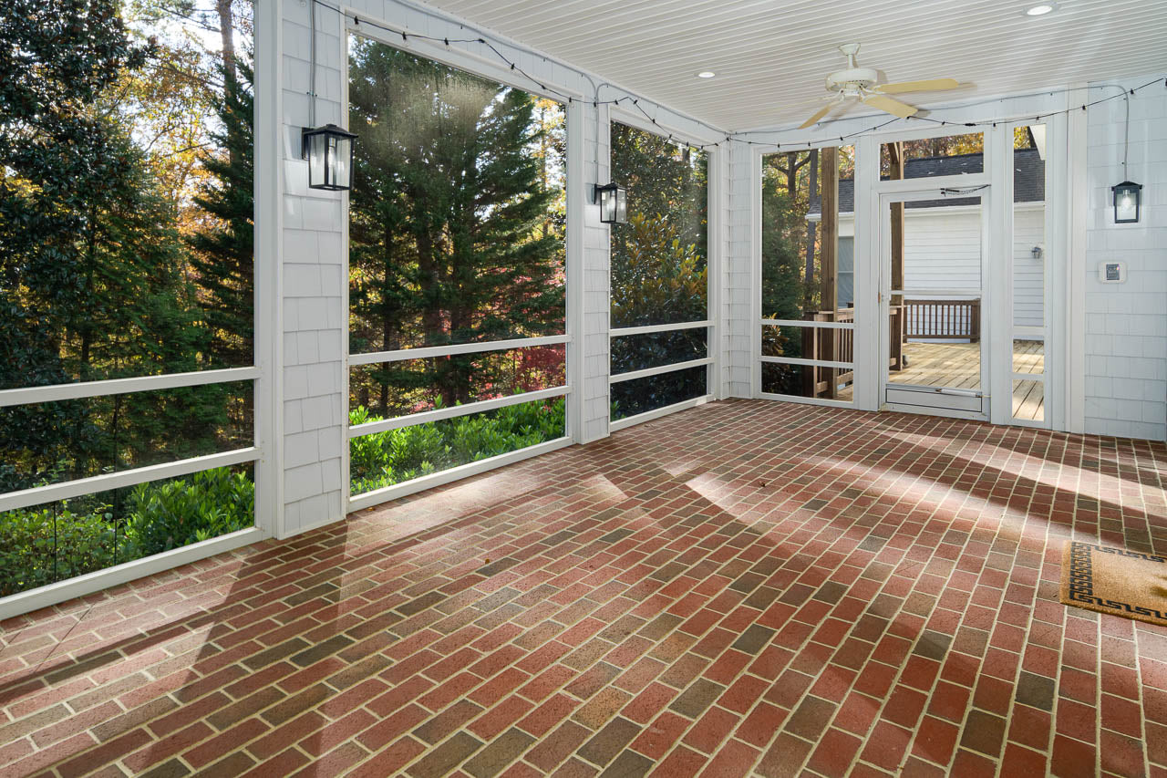 A screened-in porch with brick flooring, white walls, string lights, two black lantern-style wall lights, and a ceiling fan, overlooking a wooded area. A door leads to an open deck outside.