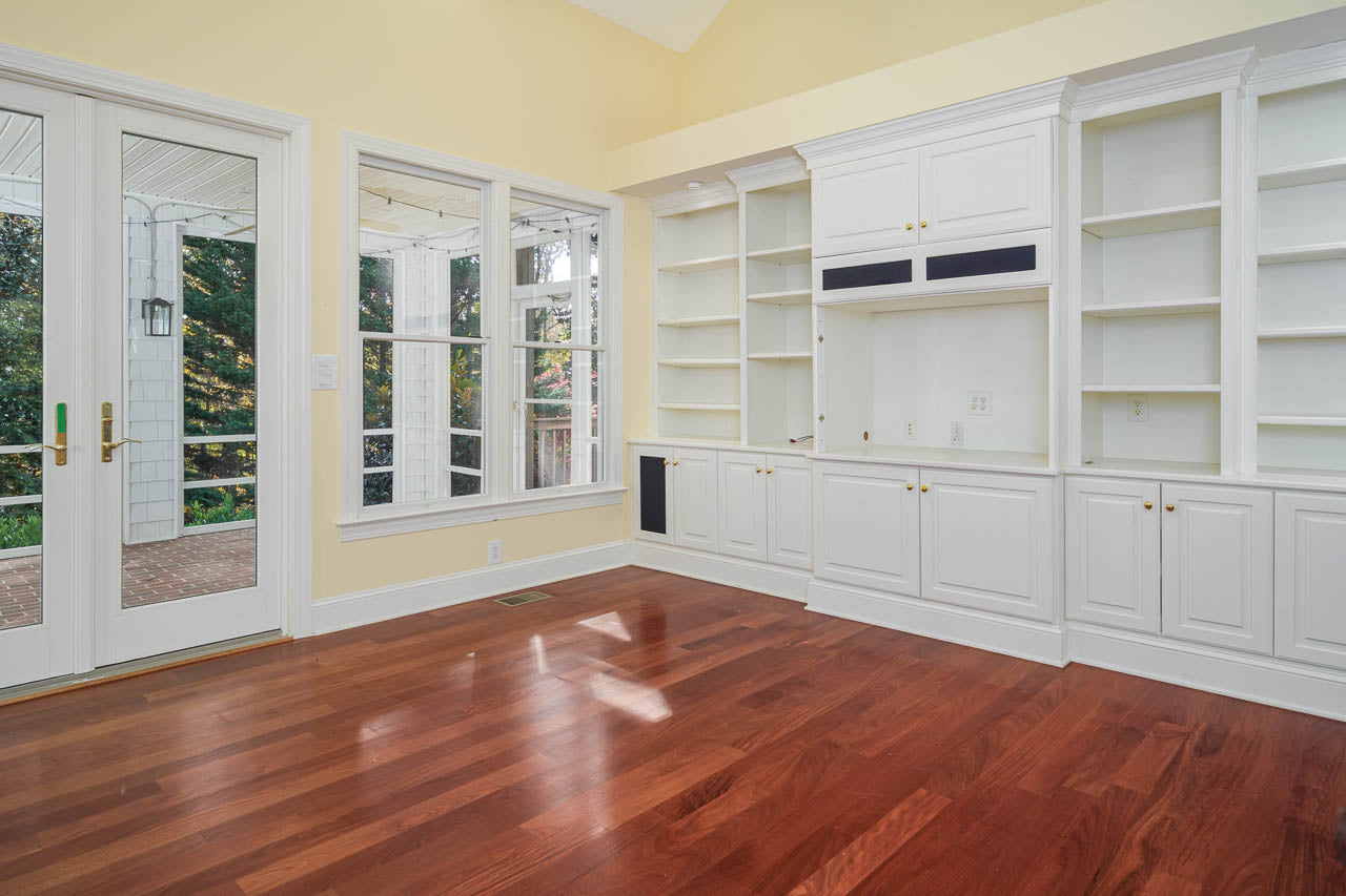 A bright room with large windows, glass doors, and built-in white shelves and cabinets. The floor is polished wood, and sunlight streams in, creating a warm, inviting atmosphere.