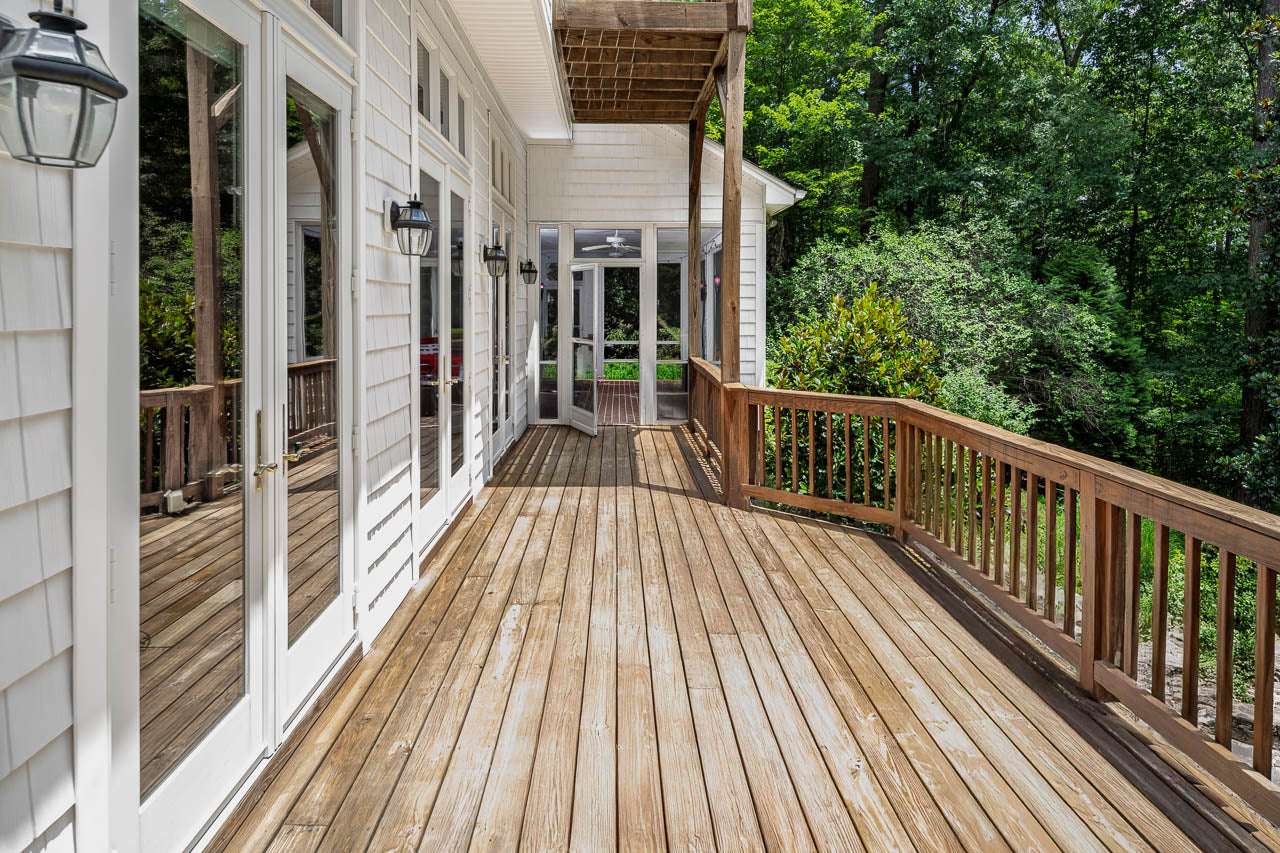 A spacious wooden deck with a wooden railing extends alongside a white house with large glass doors and windows, surrounded by green trees and foliage.