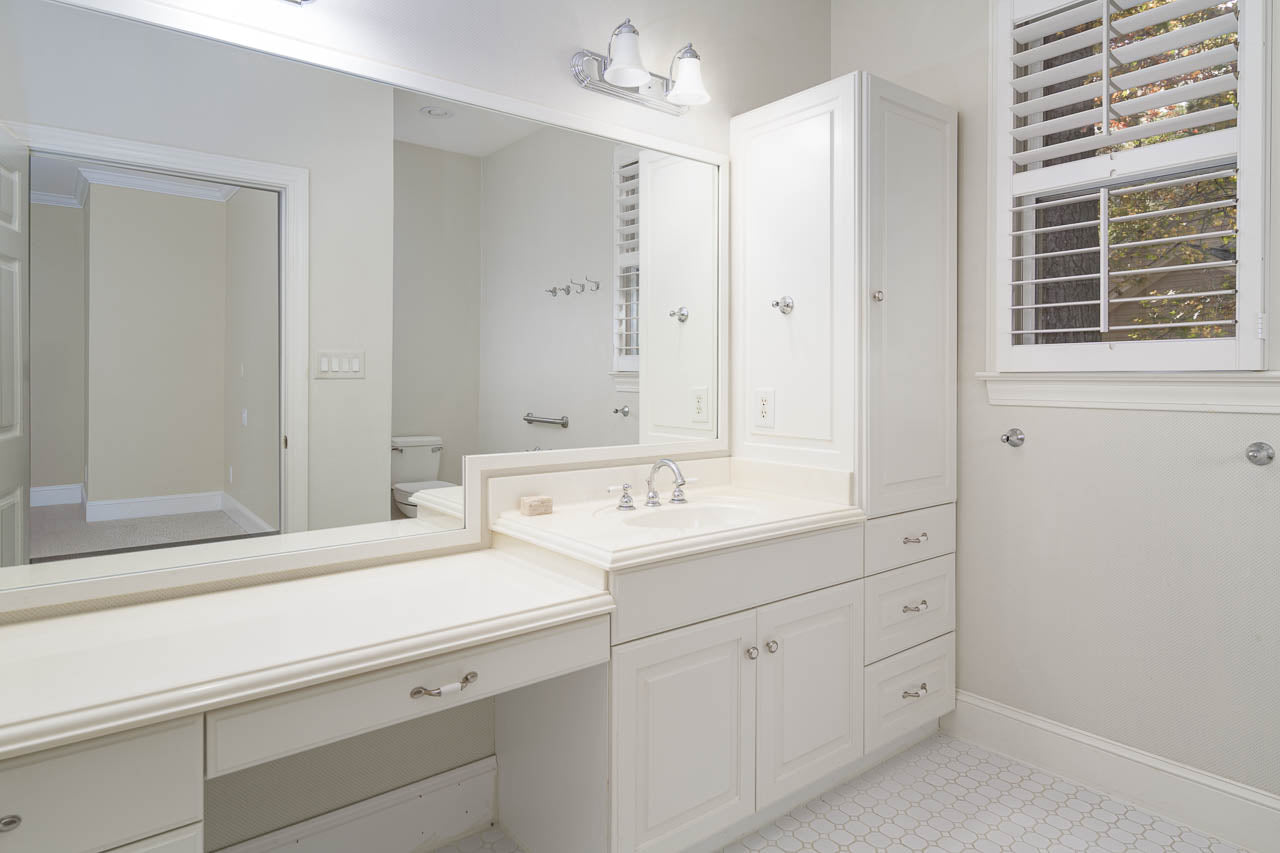 Spacious white bathroom with built-in vanity, large mirror, sink, cabinets, and drawers. A window with shutters lets in natural light. The floor has a white hexagon tile pattern.