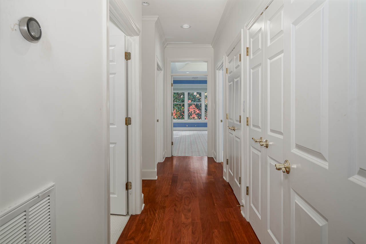 A bright hallway with white doors on both sides, wood flooring, and a view into a sunlit room with large windows and greenery outside.