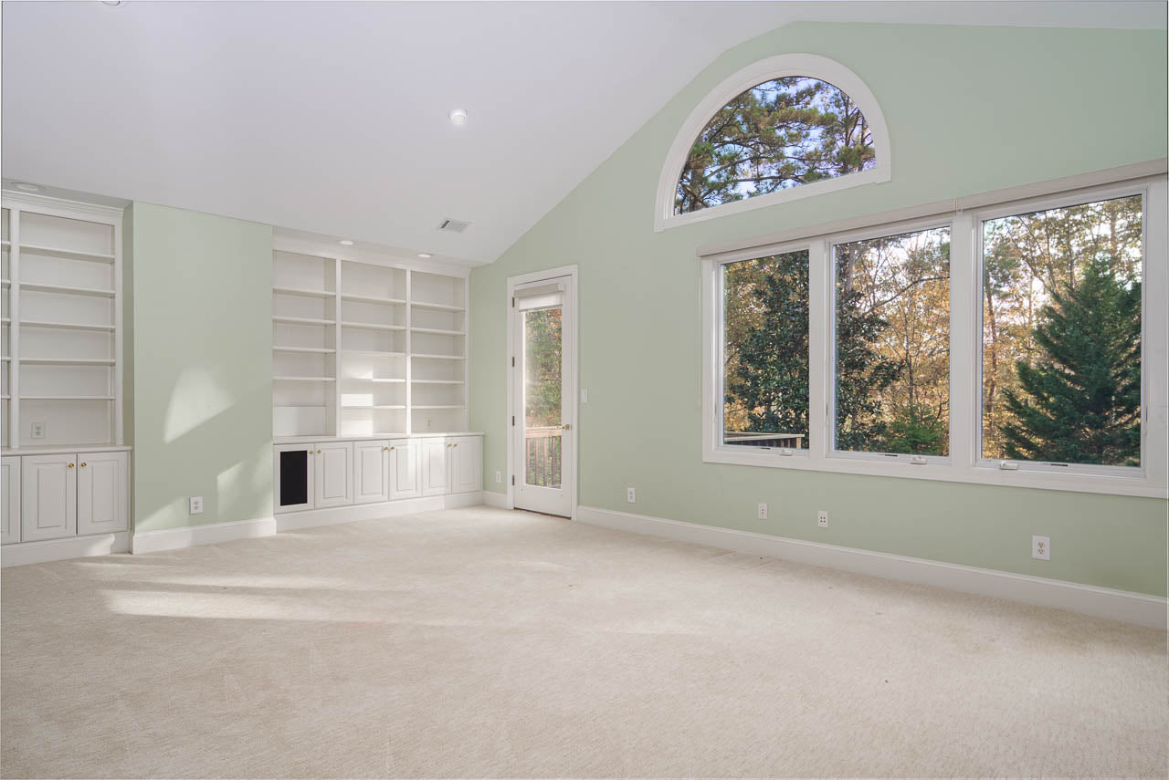 A spacious, empty room with light green walls, large windows overlooking trees, built-in white shelves and cabinets, beige carpet, and a door leading outside. Natural light fills the room.