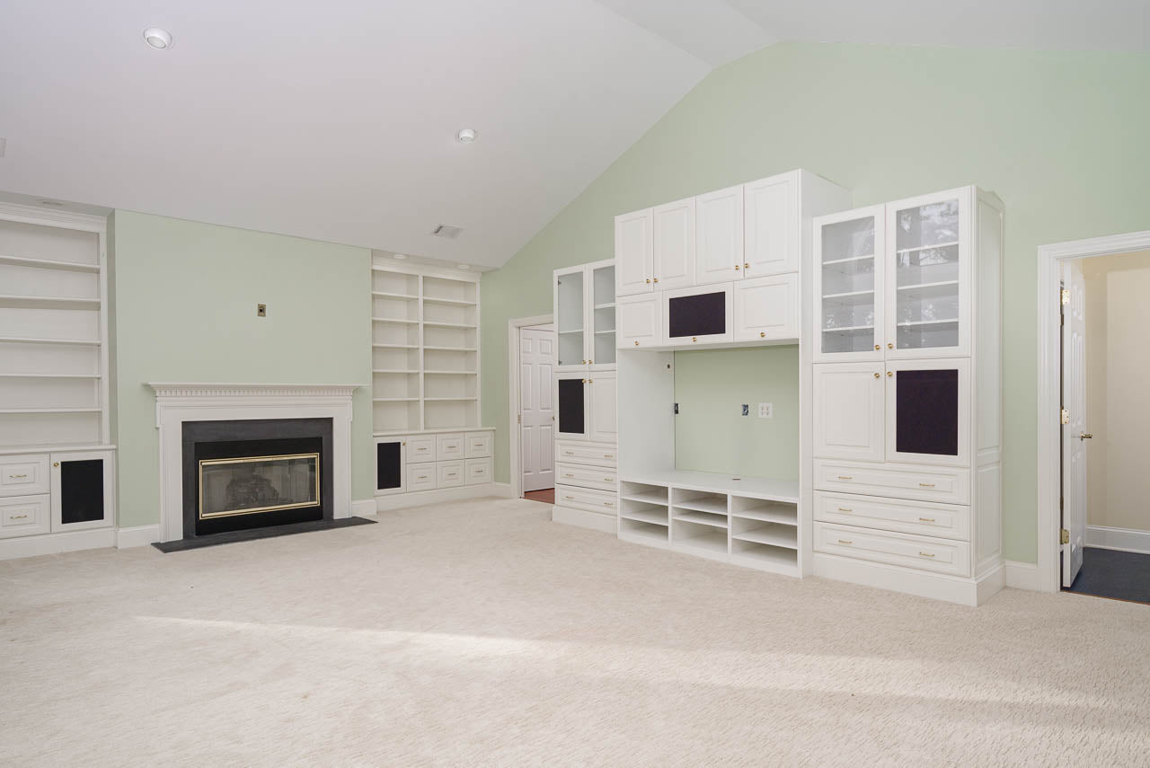 A spacious, carpeted living room with pale green walls, built-in white shelving, cabinets, and a fireplace with a black and gold trim. There is an empty entertainment center and a door to the right.