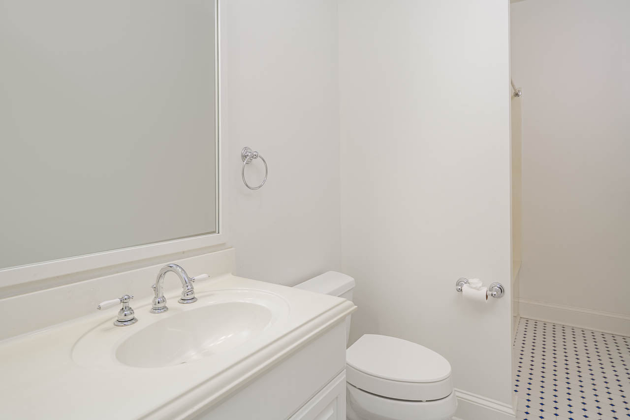 A clean, white bathroom with a sink, a silver faucet, a mirror, a toilet, a towel ring, and a toilet paper holder. The floor has a white tile pattern with small dark accents.