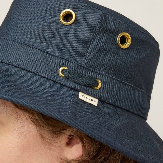 A young person with fair skin and light brown hair wears the TILLEY - ICONIC T1 BUCKET HAT IN DARK NAVY and a light gray crew-neck shirt, looking directly at the camera against a plain, light background.