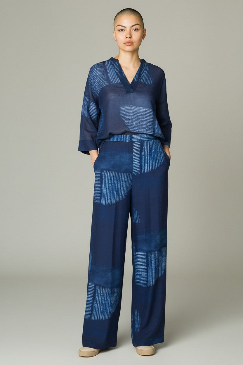 A person with a shaved head wears SANDRO FERRONE - PRINTED PANT: wide-leg, lightweight navy pants with a light blue abstract print, paired with a matching blouse, posing hands-in-pockets against a neutral background.