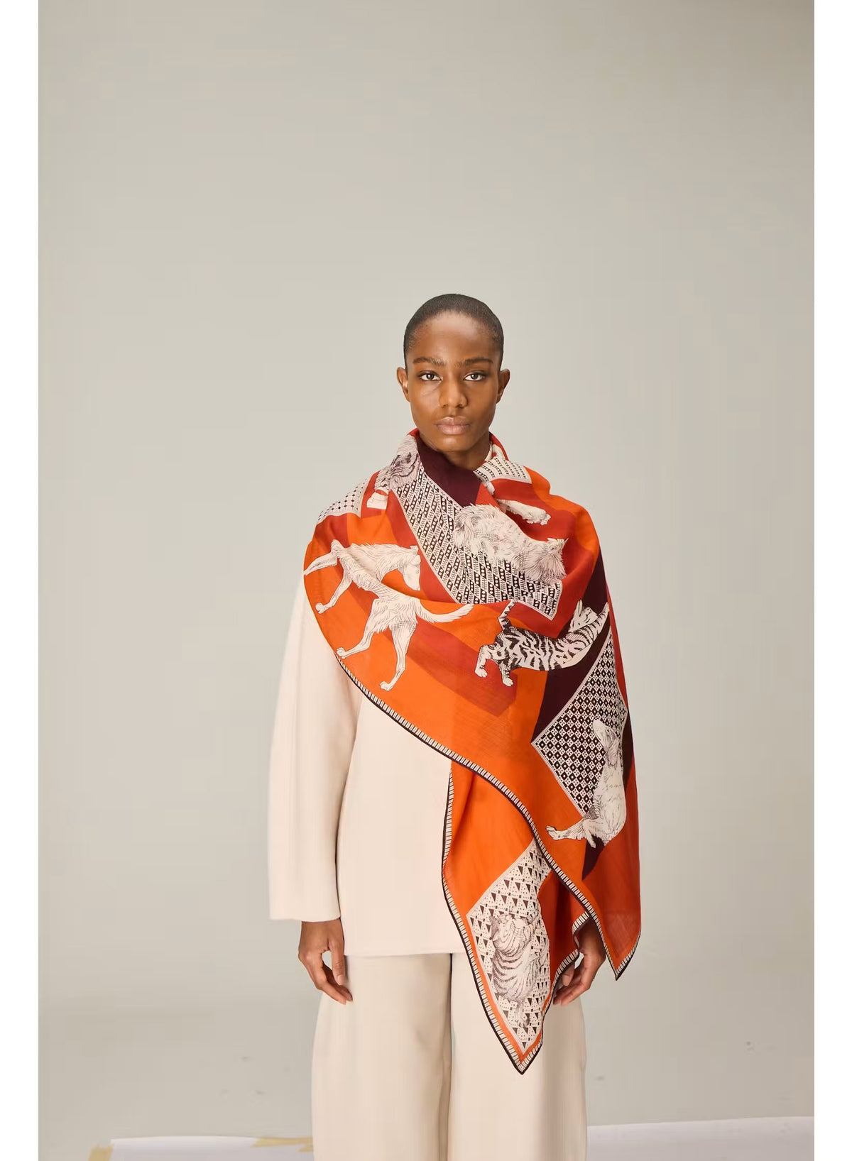A person dressed in a cream outfit models the INOUI EDITIONS - LES CHERIS WOOL SCARF IN RED by INOUI—a 100% wool accessory made in India, featuring bold orange, red, and burgundy tones adorned with large white cat illustrations and geometric patterns—set against a plain background.