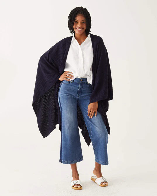 A woman with short, curly hair smiles while wearing a white blouse, wide-leg blue jeans, and the MERSEA CHARLESTON CABLE WRAP over her outfit, standing against a plain white background.