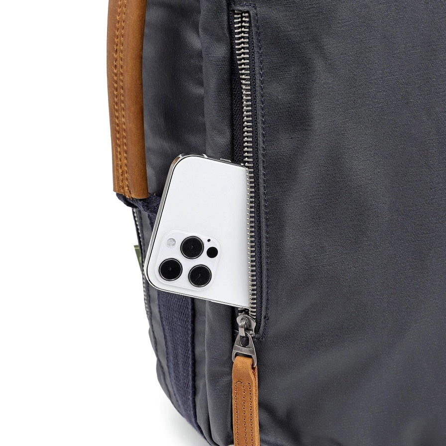 The TSD BRAND - MADRONE COATED CANVAS BACKPACK IN NAVY features brown leather accents, a spacious main compartment, a padded laptop sleeve with snap closure, and multiple zippered pockets on the inside flap.