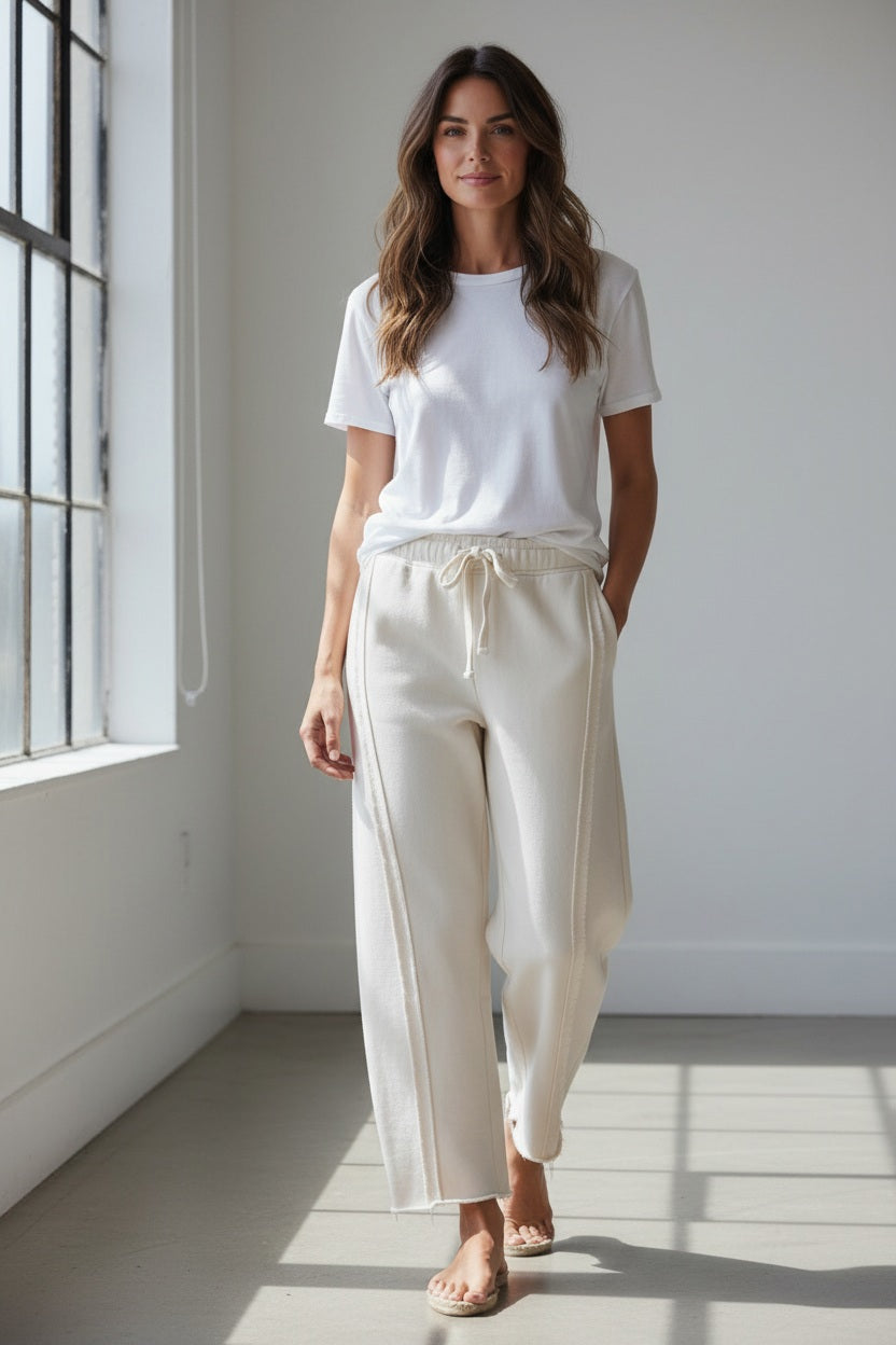A person stands in a doorway wearing the FRANK & EILEEN Bibi Barrel Leg Sweatpant in Triple Fleece Vintage White, featuring raw edge side seams. Only the lower half of their body is visible. They pair the sweatpants with a striped top and tan boat shoes, with one hand resting on their hip.
