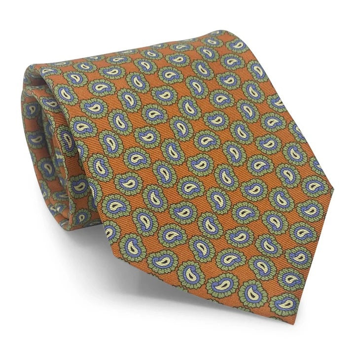 Introducing the PAGE SILK TIE IN ORANGE by BIRD DOG BAY. This expertly handmade New York tie features a vibrant orange base adorned with a repeating blue and yellow paisley pattern edged in green. With its pointed tip and solid white background, the PETER-BLAIR tie elevates classic design for a modern, sophisticated look.