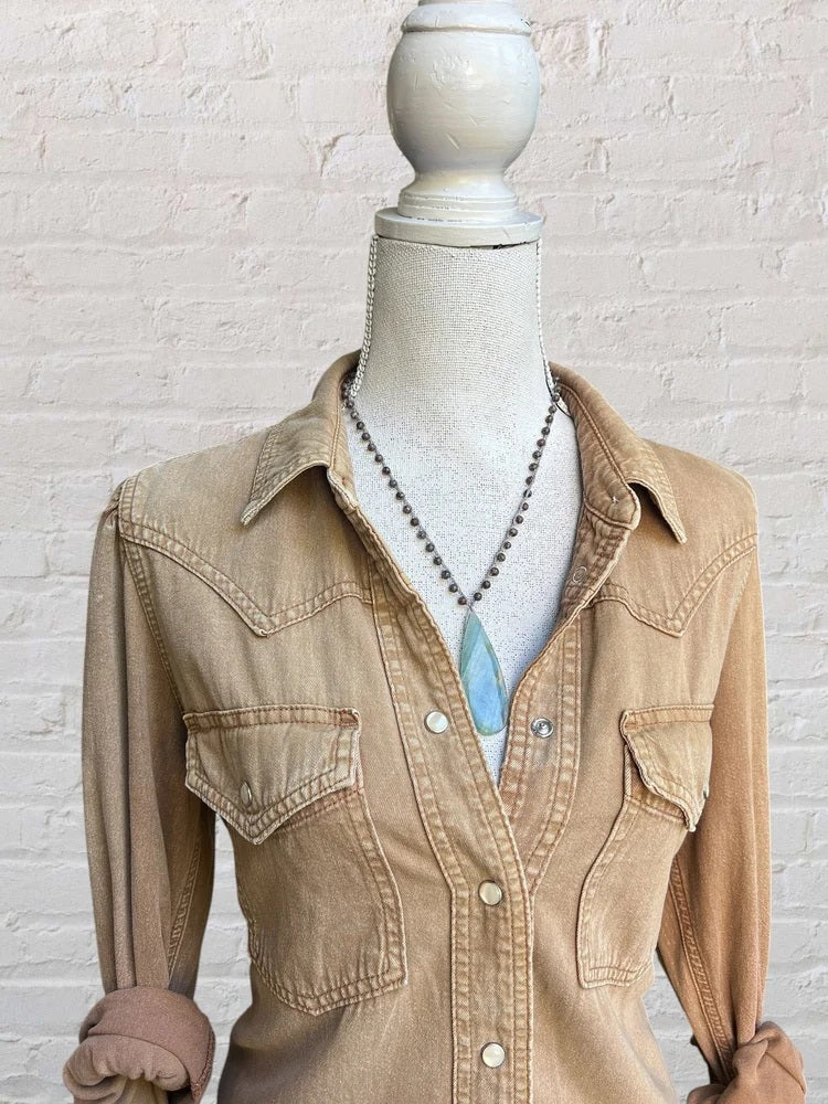 A mannequin dressed in a light brown button-up shirt with rolled sleeves and front pockets, wearing the ANN LIGHTFOOT Blue Calcite Drop Pendant and Snowflake Jasper Necklace, stands in front of a white brick wall.