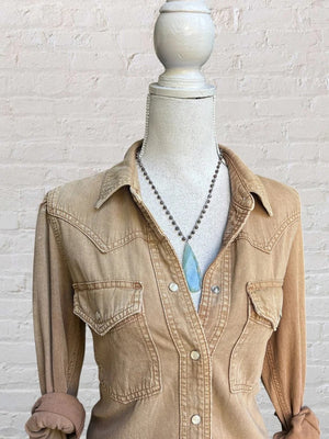 A mannequin dressed in a light brown button-up shirt with rolled sleeves and front pockets, wearing the ANN LIGHTFOOT Blue Calcite Drop Pendant and Snowflake Jasper Necklace, stands in front of a white brick wall.