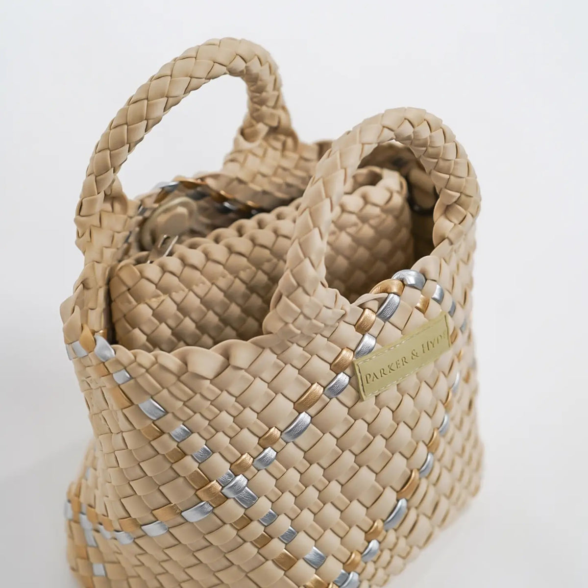 A woman with long brown hair, in a sleeveless cream knit top and blue jeans, holds a PARKER AND HYDE - MINI WOVEN TOTE BAG. The hand-woven tote by PARKER &amp; HYDE features a subtle checkered pattern with light blue accents.