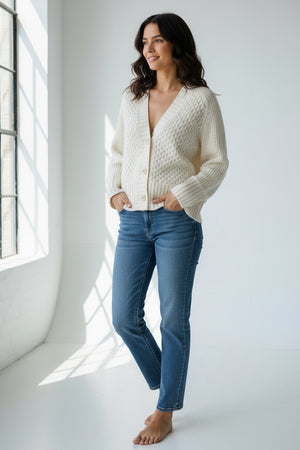 A person models the KINROSS CASHMERE Luxe Mixed Stitch Cardigan in cream, featuring large buttons and textured 8ply knit, paired with blue jeans, hands in pockets, facing the camera against a plain white background.