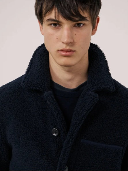 A young person with short dark hair wears the HARTFORD - Sherpa Utility Jacket in Navy by HARTFORD over a black shirt, looking directly at the camera against a plain light background. This warm sherpa layer brings both comfort and style to casual menswear.