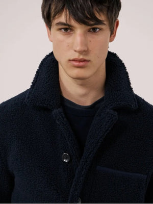 A young person with short dark hair wears the HARTFORD - Sherpa Utility Jacket in Navy by HARTFORD over a black shirt, looking directly at the camera against a plain light background. This warm sherpa layer brings both comfort and style to casual menswear.