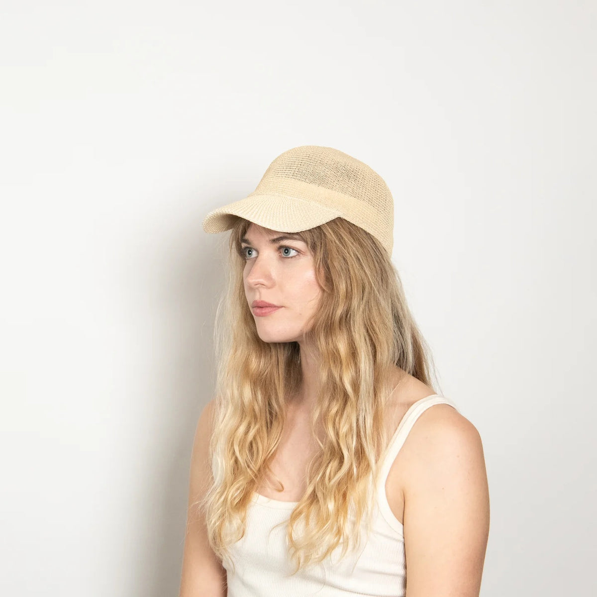 A woman with long, wavy blonde hair wears the WYETH HATS - PAULA MESH CAP IN NATURAL by WYETH and a white tank top, sitting against a plain light background and looking to the left with a neutral expression.