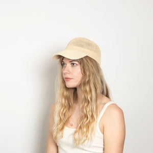 A woman with long, wavy blonde hair wears the WYETH HATS - PAULA MESH CAP IN NATURAL by WYETH and a white tank top, sitting against a plain light background and looking to the left with a neutral expression.