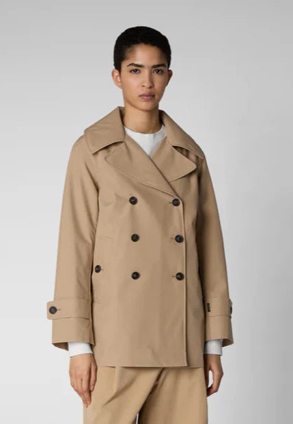 An individual with short blonde hair is wearing the SAVE THE DUCK - SOFI TRENCH JACKET, a beige, weather-resistant double-breasted coat. They are also dressed in a white top, black pants, and black pointed shoes. They are standing against a plain gray background, facing forward with arms at their sides.