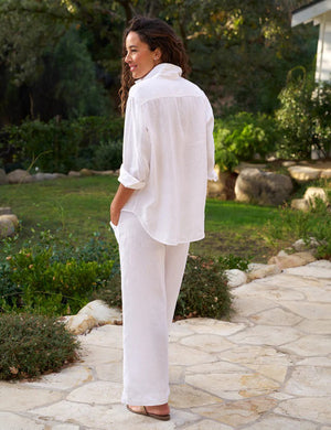 A woman with curly hair smiles outdoors, wearing the FRANK & EILEEN - EILEEN Relaxed Button Up Shirt in linen from the Tuscany Getaway Set, paired with wide-leg pants, standing on a sunlit stone patio surrounded by greenery.