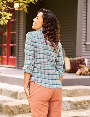 A woman with curly hair smiles and looks up as she stands outside next to a tree, wearing the Frank & Eileen Barry Tailored Button Up in Cream Green Blue Plaid Italian Flannel and rust-colored pants. Her hands are in her pockets, and a house with a red door and a pile of firewood is visible in the background.