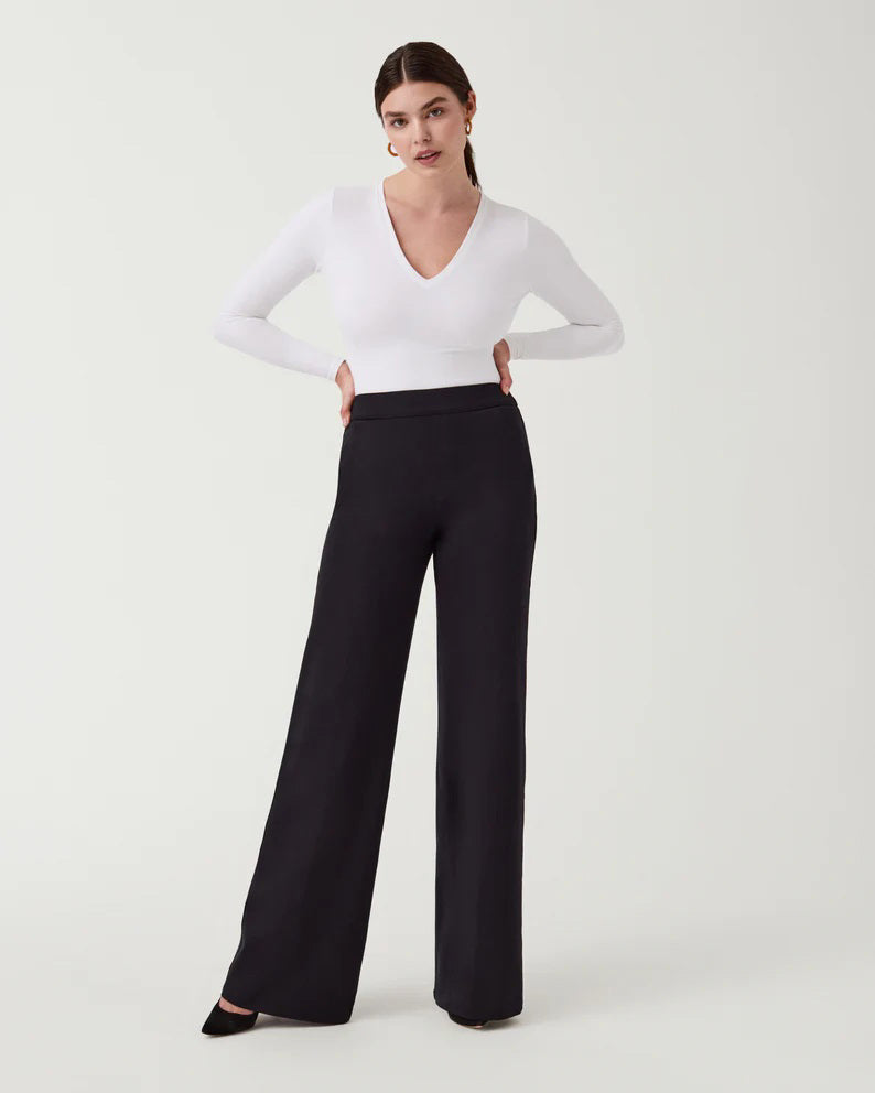 A woman stands against a plain background wearing the SPANX Perfect Pant Wide Leg in black—crafted from premium ponte fabric—paired with a white long-sleeve V-neck top and black heeled shoes.